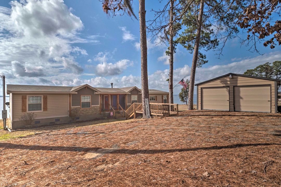 Single-story house with wooden accents, surrounded by tall pine trees, beside a large garage. An American flag waves in the breeze under a partly cloudy sky.