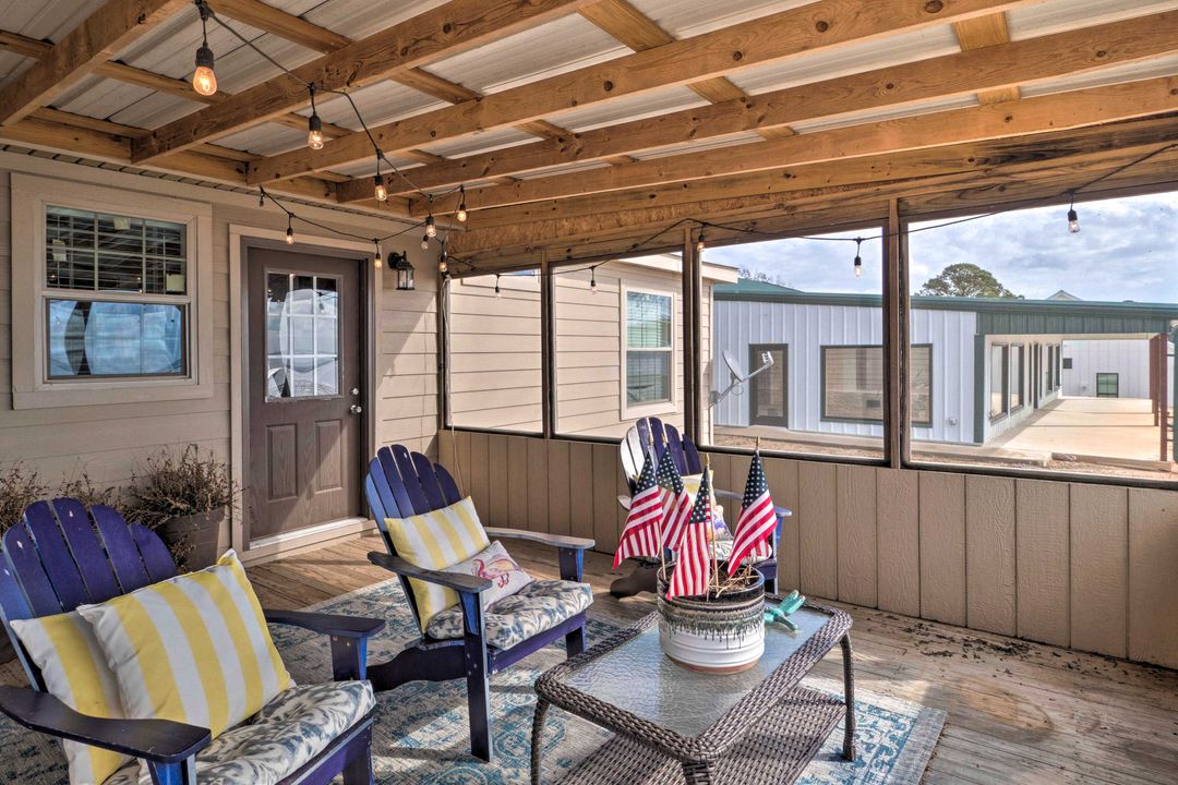 Cozy porch with wooden beams, string lights, blue chairs with yellow striped cushions. Small table holds American flags. Calm, inviting atmosphere.