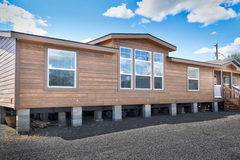 A brown manufactured home elevated on concrete blocks with large windows, set against a clear blue sky and white clouds, creating a bright, serene atmosphere.