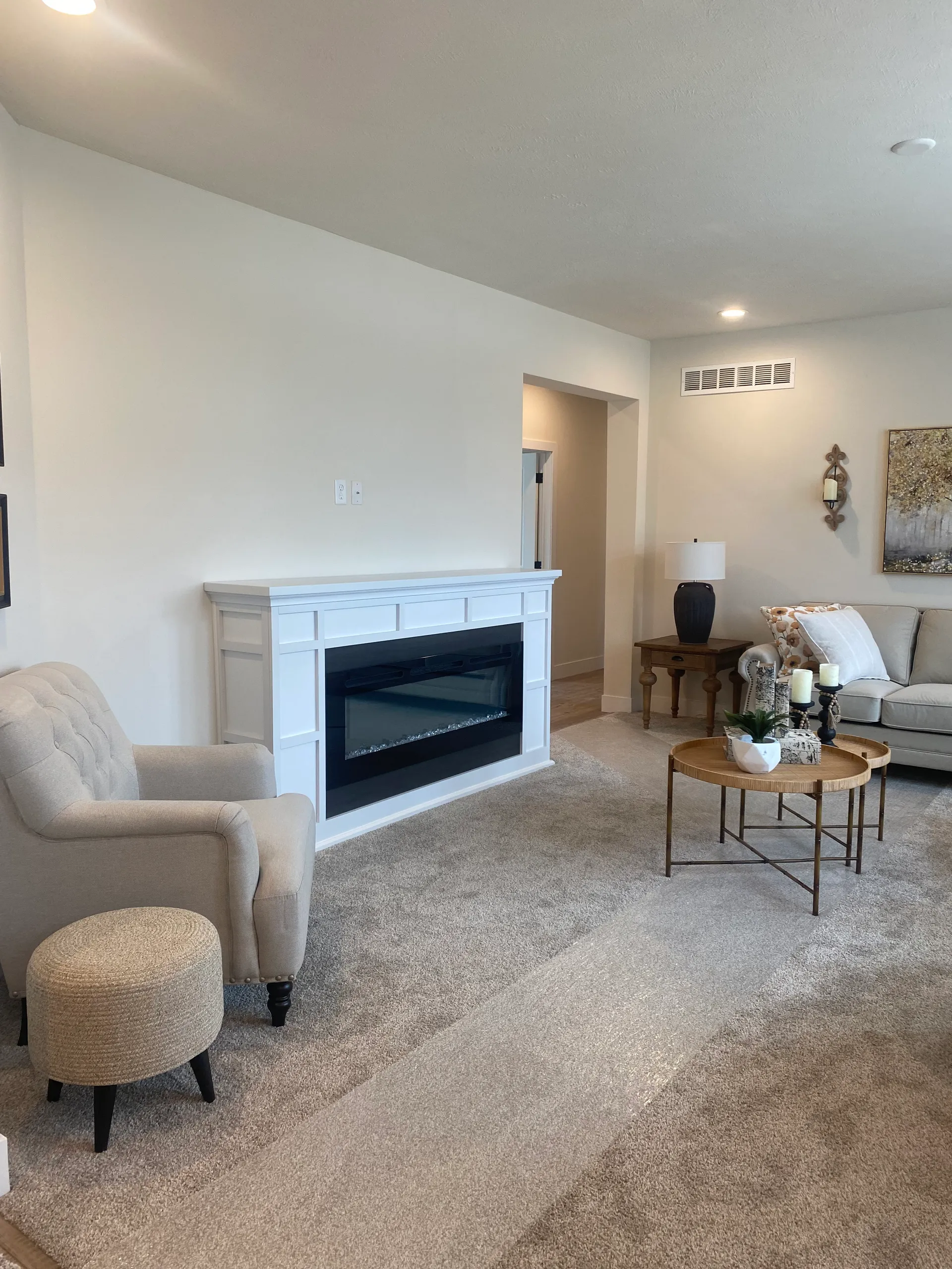 Cozy living room with beige carpet and light walls. Features a white fireplace, beige armchair, round wooden table, and a sofa with patterned cushions. Warm, inviting atmosphere.