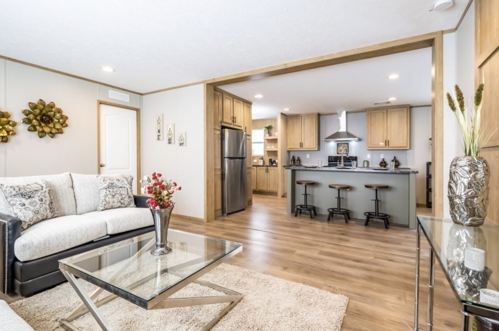 Bright open-plan living room with modern kitchen. Cream sofa, glass coffee table, and bar stools. Light wood floors and cabinets. Cozy, inviting ambiance.