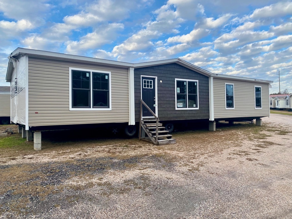 A beige modular home with black accents sits on a gravel lot under a partly cloudy blue sky. It features large windows and a small staircase leading to a black door.