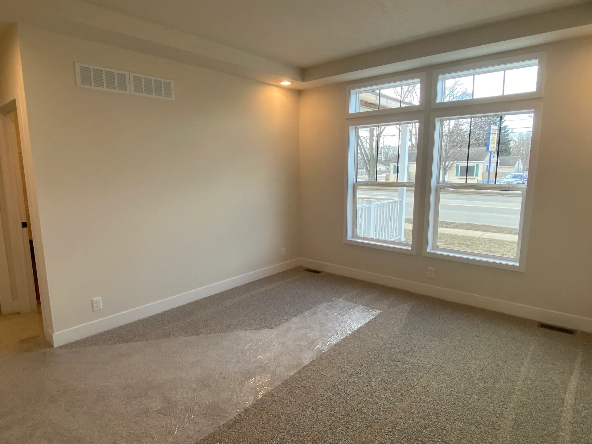 Empty room with beige walls and carpet, illuminated by soft recessed lighting. Large windows with white trim offer a clear view of the street outside. Peaceful and bright ambiance.
