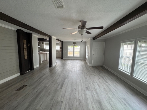 Spacious empty room with gray wood floors, white walls, and dark ceiling beams. Features a ceiling fan, large windows, and abundant natural light.
