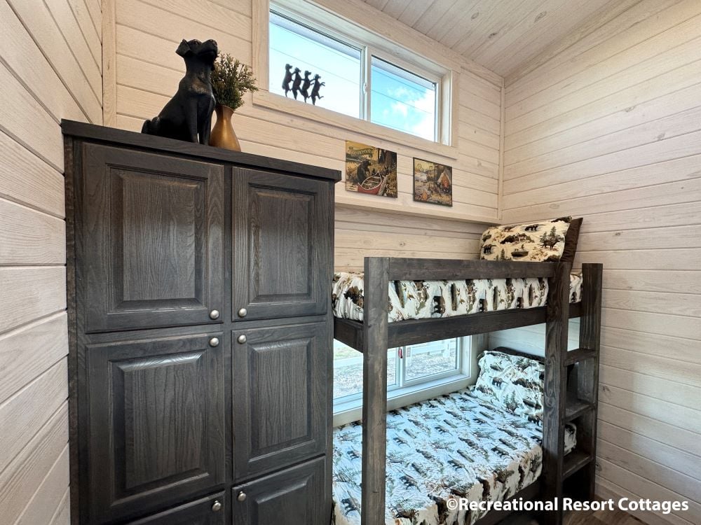 Cozy cabin room with wooden walls, featuring a bunk bed with rustic-patterned bedding, a tall dark wood cabinet, and small western-style artworks.