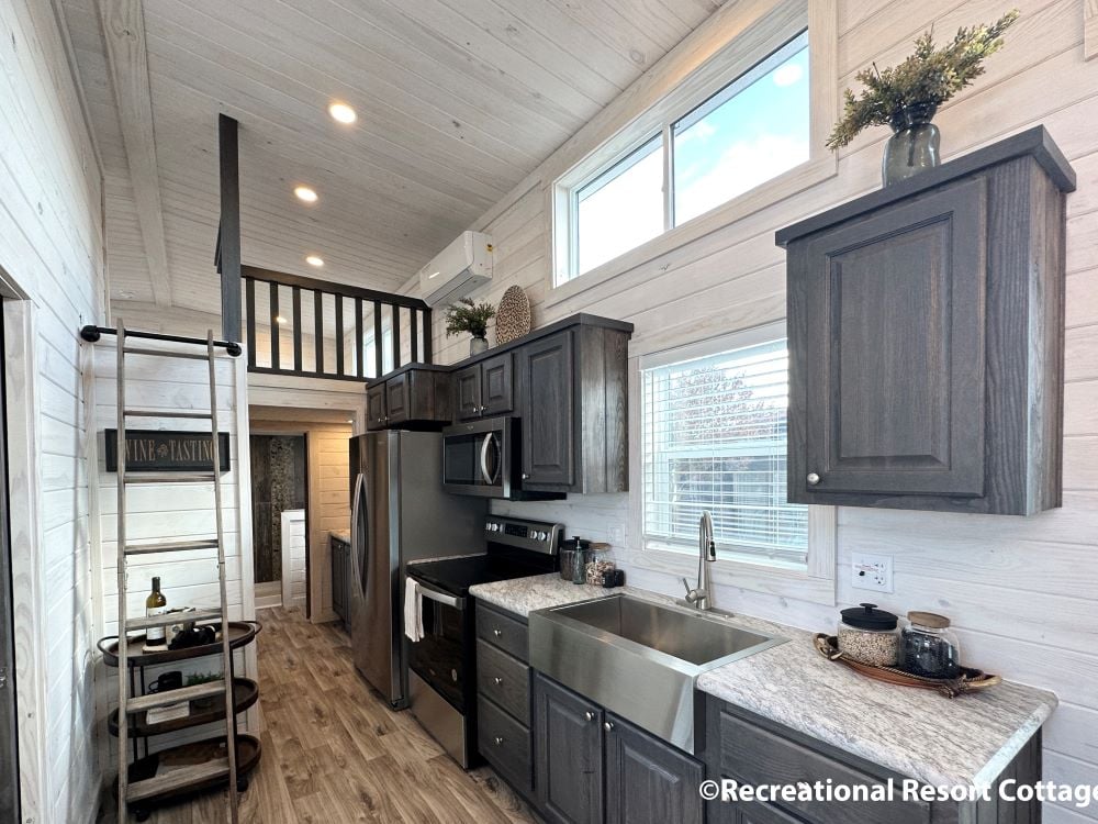 Cozy tiny house kitchen with rustic wooden cabinets, stainless steel appliances, and a loft accessed by a ladder. Natural light from large windows.