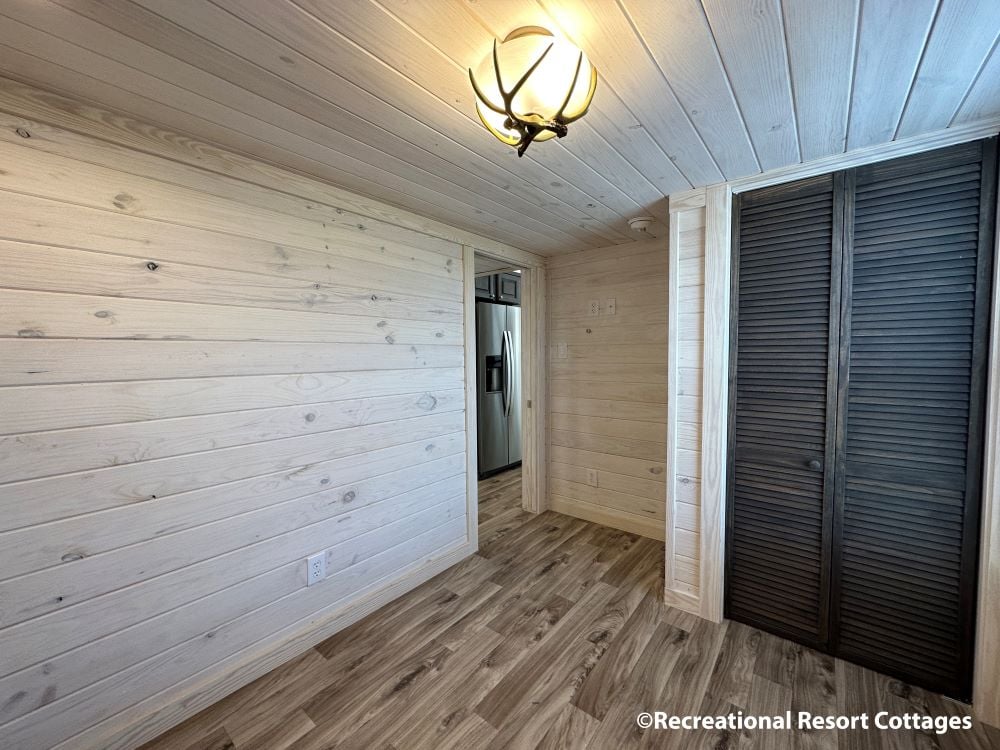 Cozy room with light wood paneling and flooring. A black louvered closet door is on the right, with a chrome fridge visible through a doorway. A soft-lit ceiling light adds warmth.