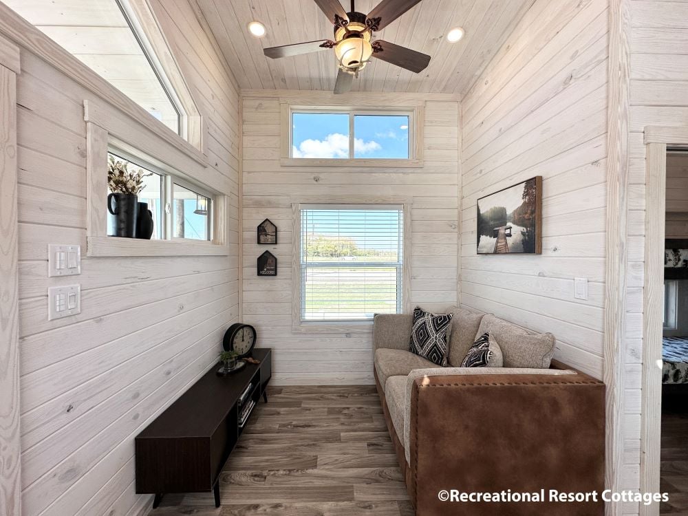 Cozy room with white wooden walls, brown couch, and decorative pillows. A small window and ceiling fan brighten the space, exuding warmth.