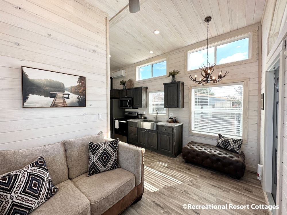 Bright, modern cottage living area with light wood paneling. Features cozy sofa, geometric pillows, rustic kitchen, large windows, and antler chandelier.