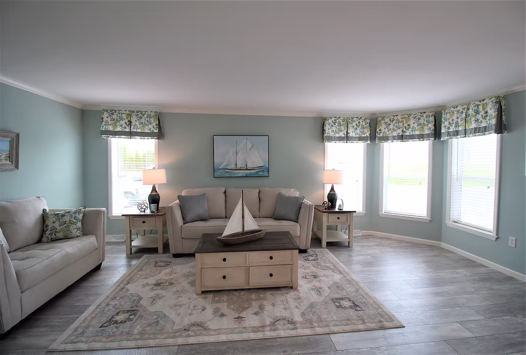 Living room with pale blue walls and wooden floor. Beige sofas and a table with a sailboat model create a nautical theme. Bright windows enhance the airy feel.