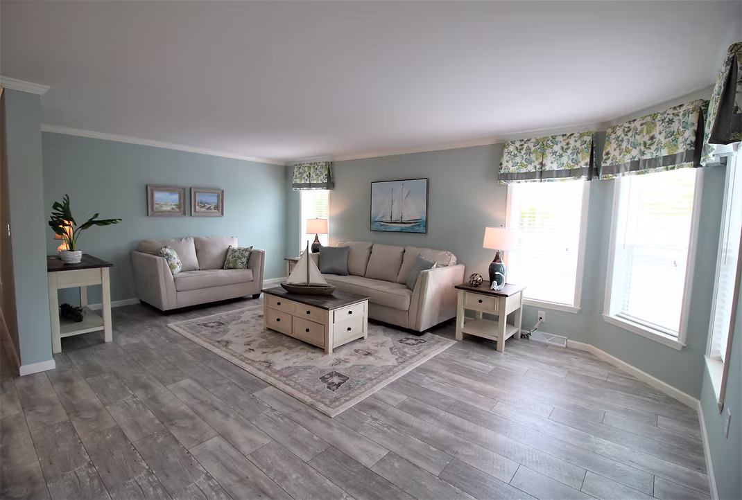 Spacious living room with pastel blue walls, light gray sofas, nautical decor, and large windows with floral curtains. Warm, inviting atmosphere.