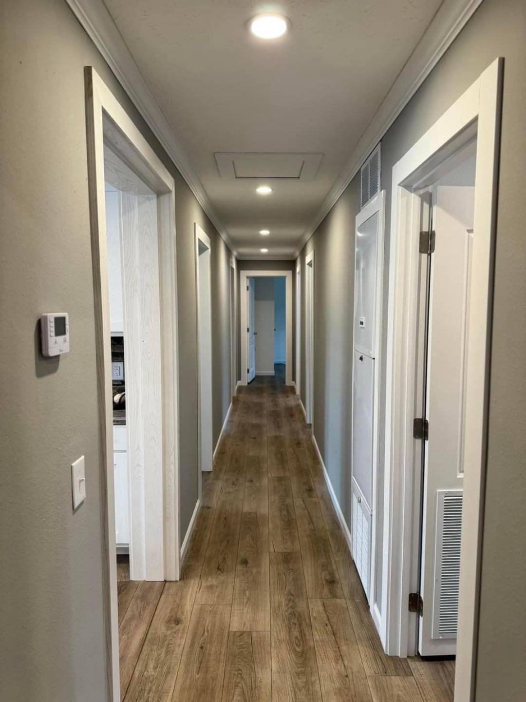 Well-lit, narrow hallway with wooden flooring, gray walls, white trim, and several doors. The atmosphere is clean and orderly.