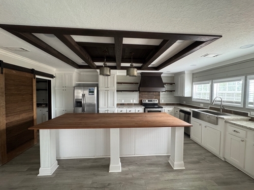 Modern kitchen with a large wooden island, white cabinets, stainless steel appliances, and a dark wood beam ceiling. Bright light from multiple windows.