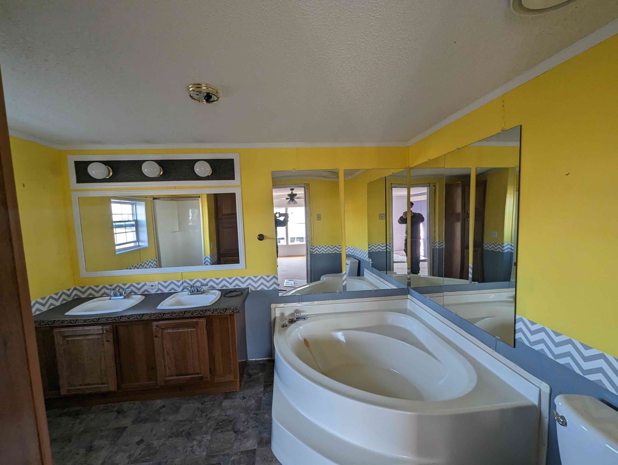 Bright bathroom with yellow walls, chevron accents, and large mirrors. Features a double sink vanity with wooden cabinets and a corner bathtub.