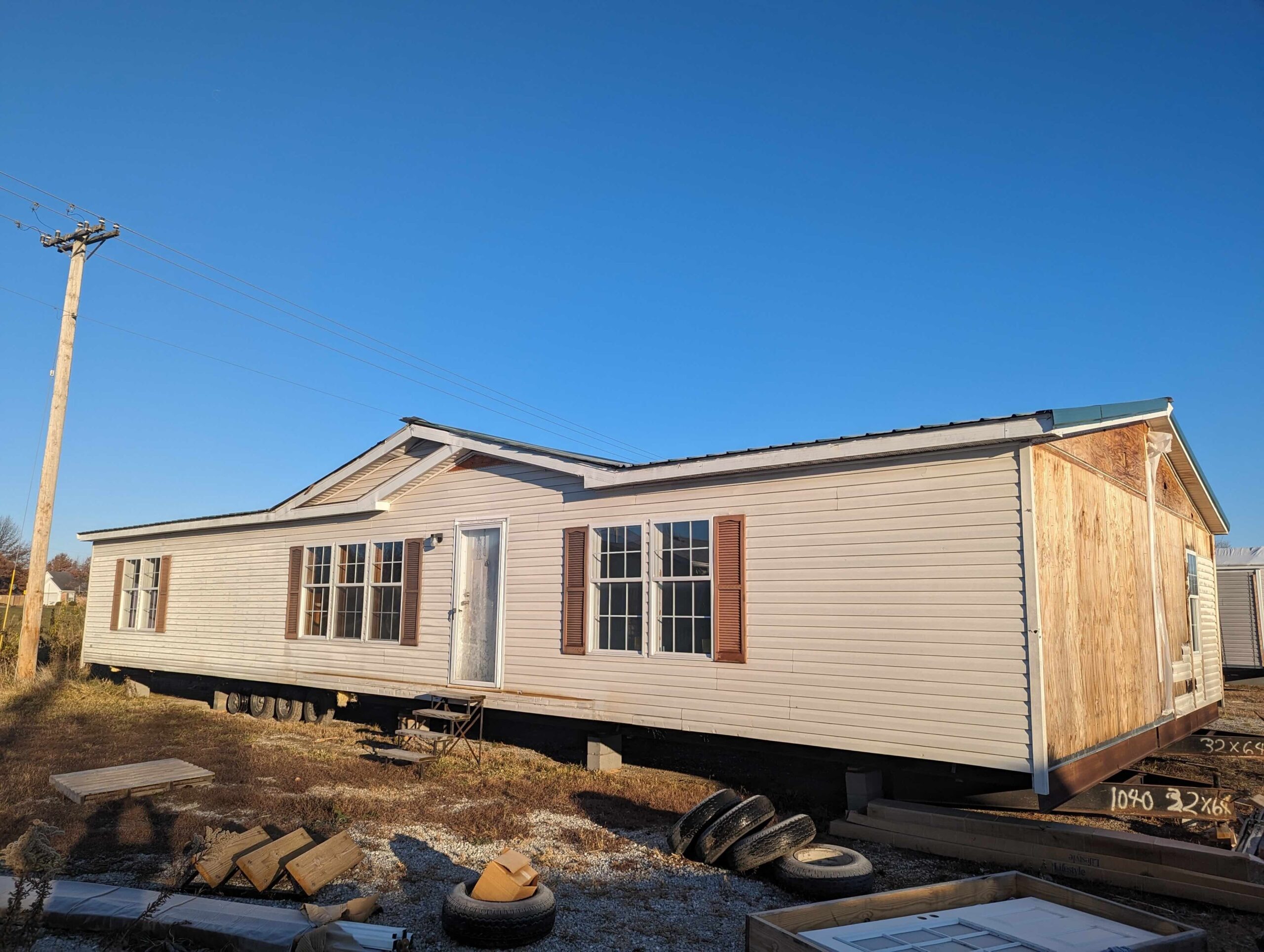 A beige mobile home on a lot, with brown shutters and multiple windows. The sky is clear blue, and tires are scattered in the foreground, conveying a peaceful, rustic setting.
