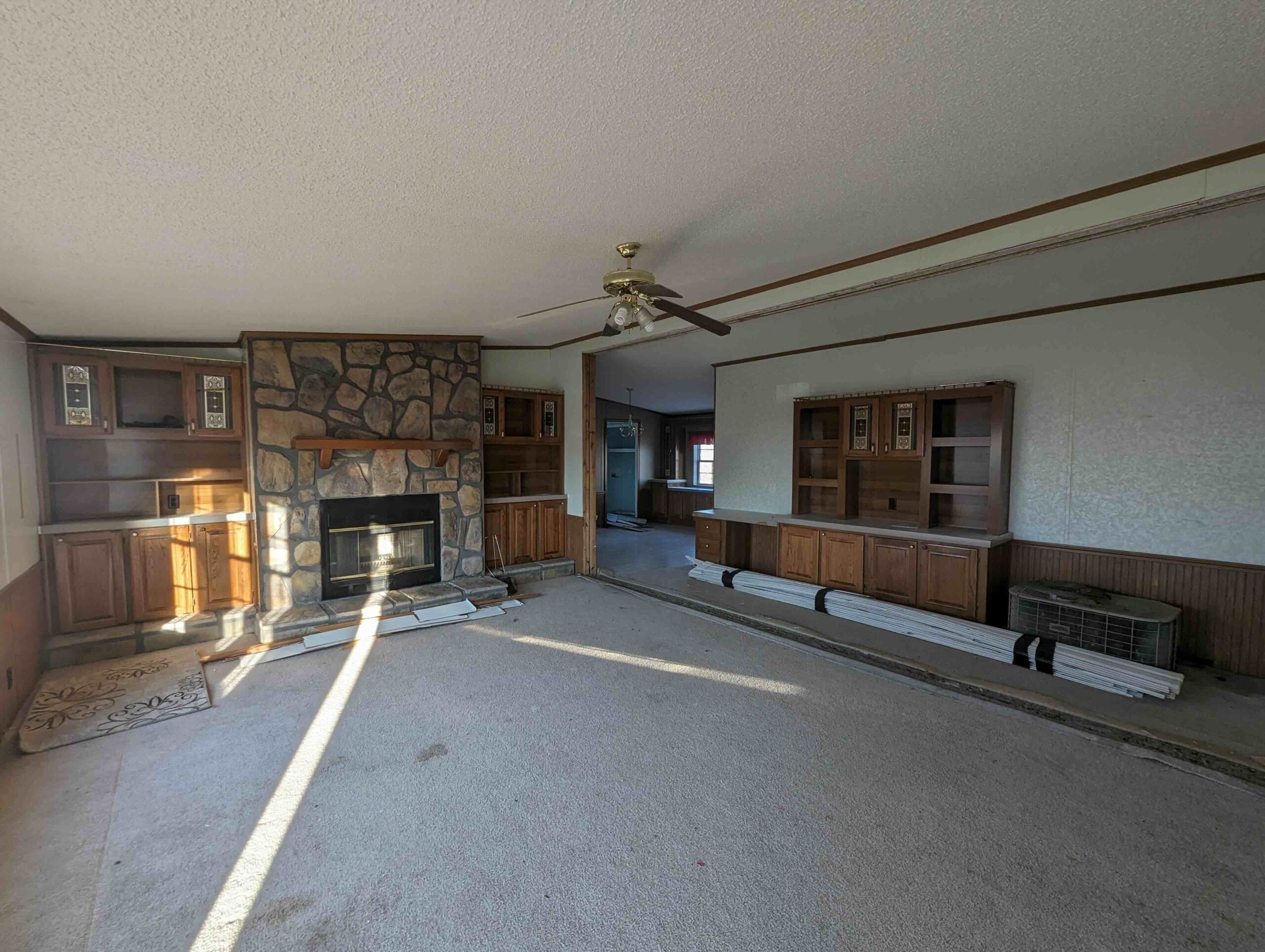 Spacious living room with a stone fireplace and wood cabinets. Ceiling fan, neutral carpet, and natural light create a calm, inviting ambiance.