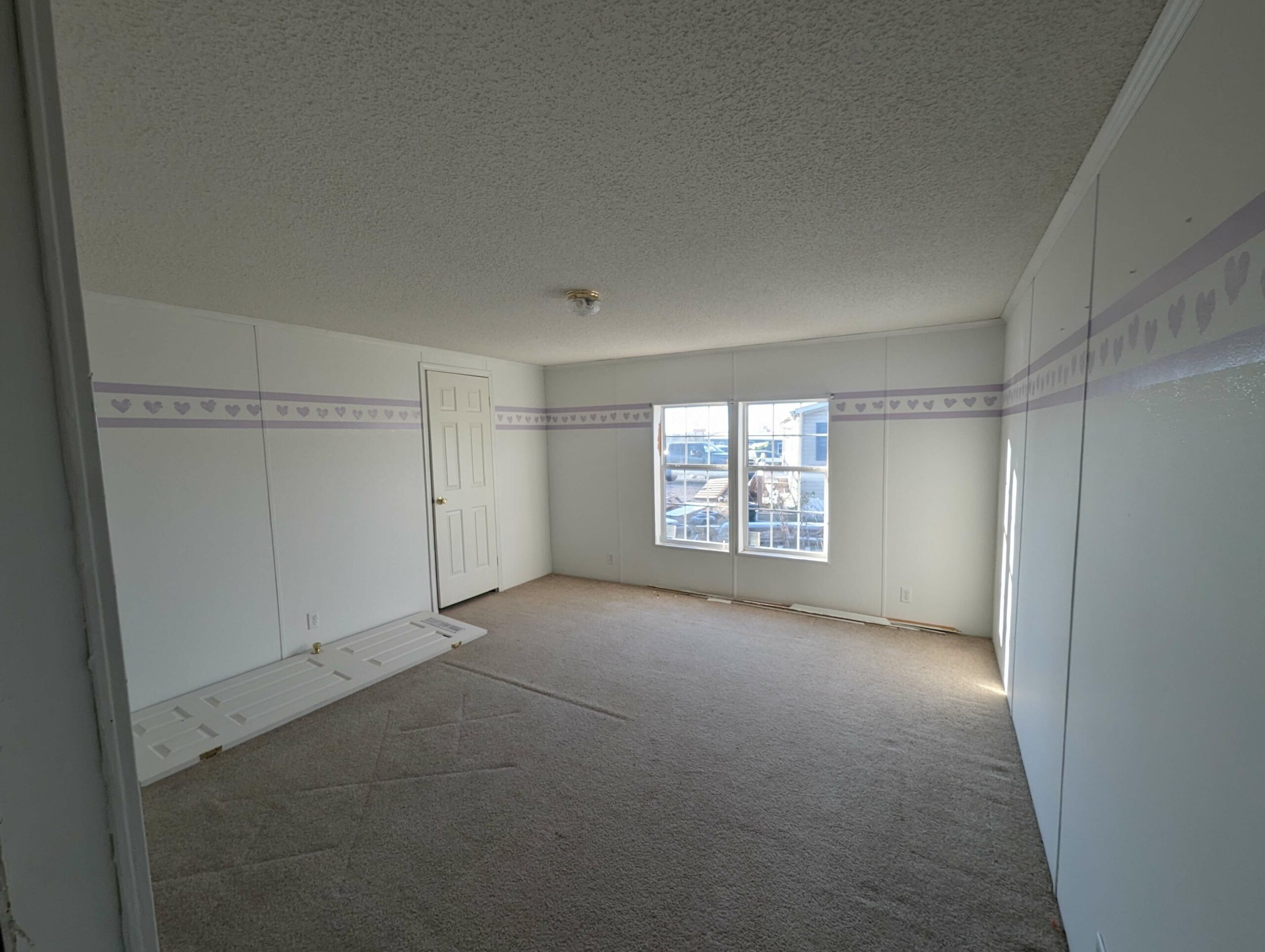 Empty room with white walls and carpet, featuring a border of purple hearts. Sunlight streams through a large window, creating a calm, inviting atmosphere.