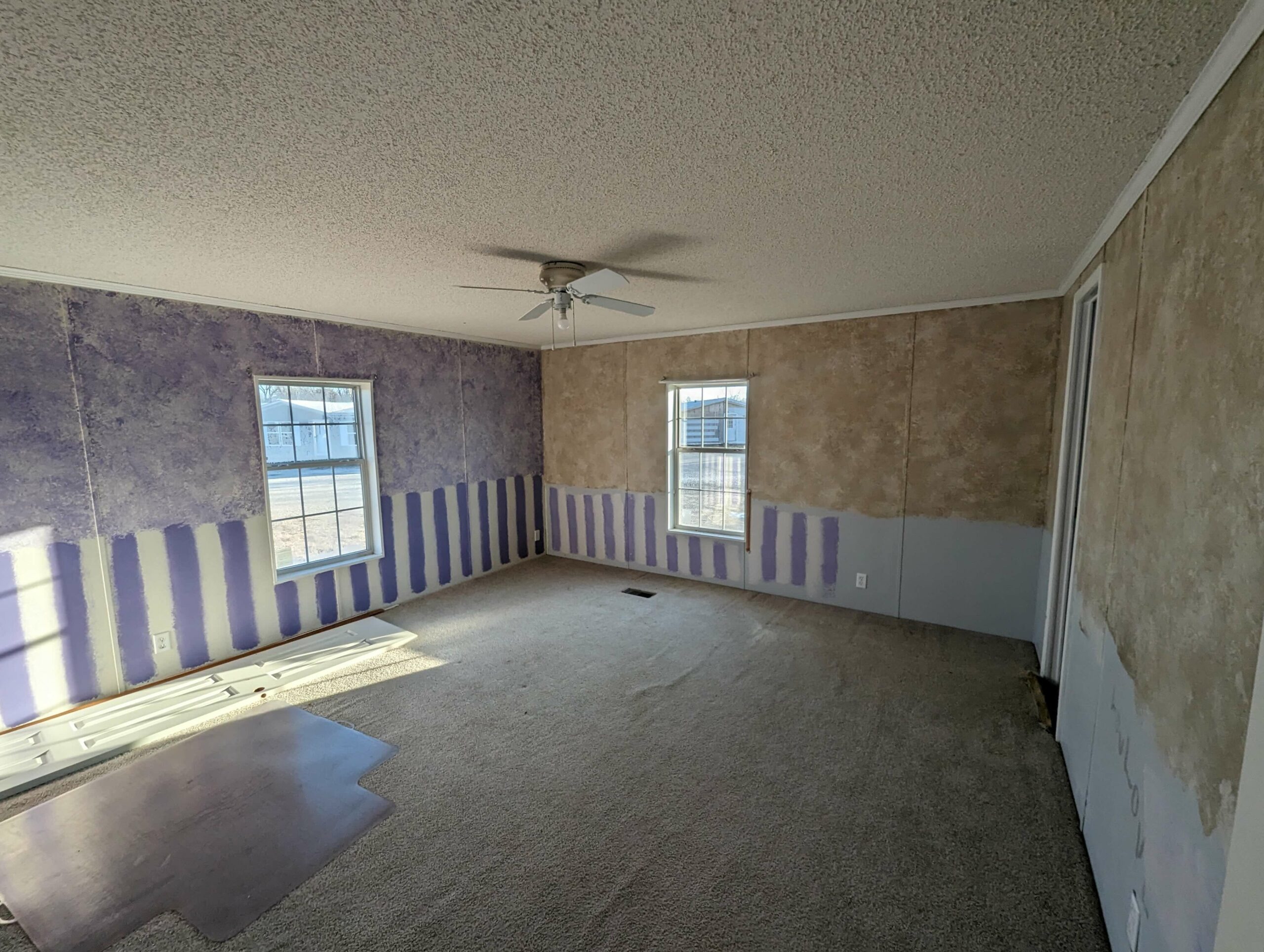 Empty room with textured purple and beige walls, featuring vertical stripes. Two windows, a ceiling fan, and beige carpet create a calm, unfinished atmosphere.