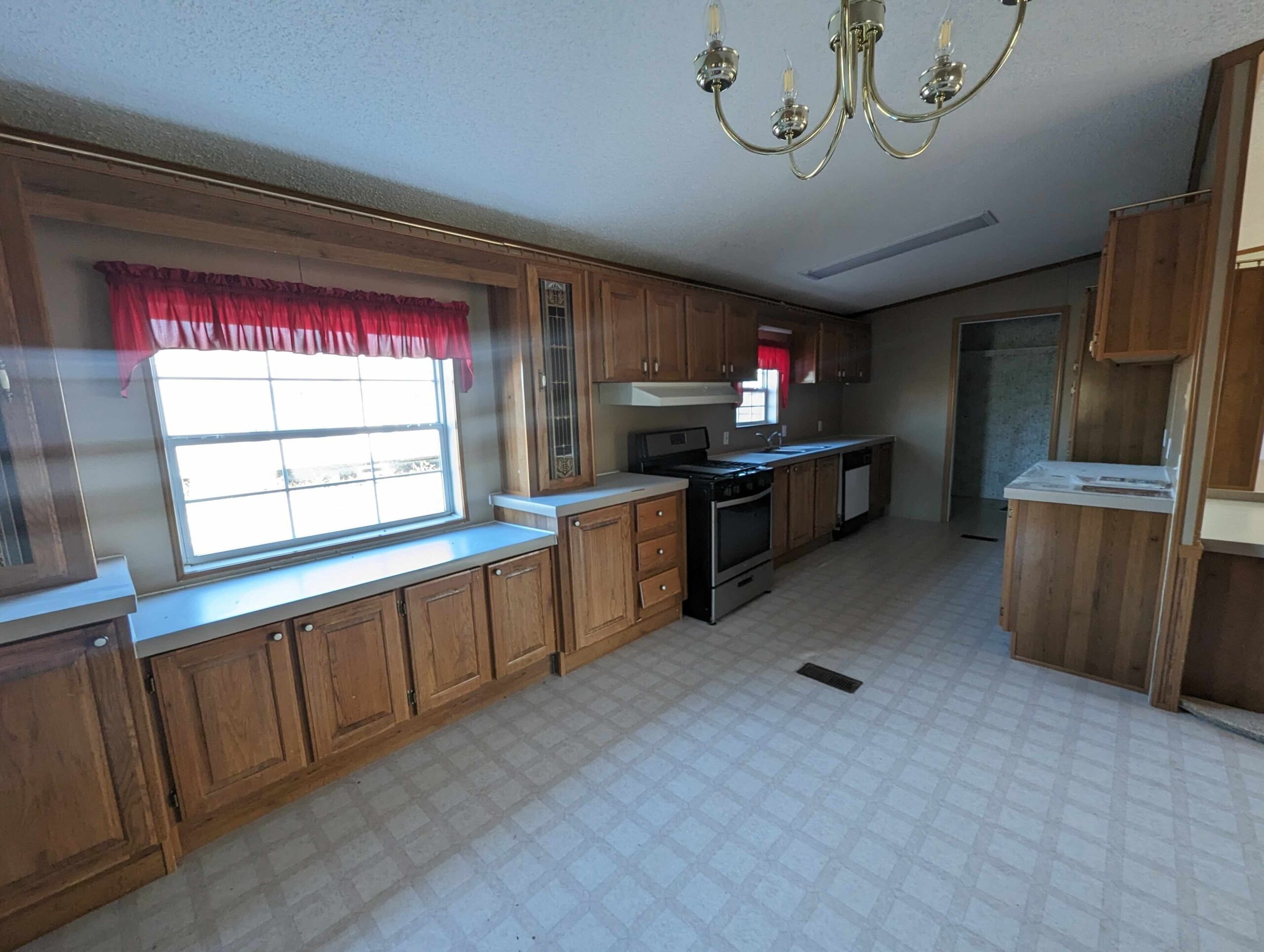 Spacious kitchen with wooden cabinets, stainless steel appliances, and large windows with red curtains. A chandelier hangs from the ceiling, creating a warm, welcoming atmosphere.