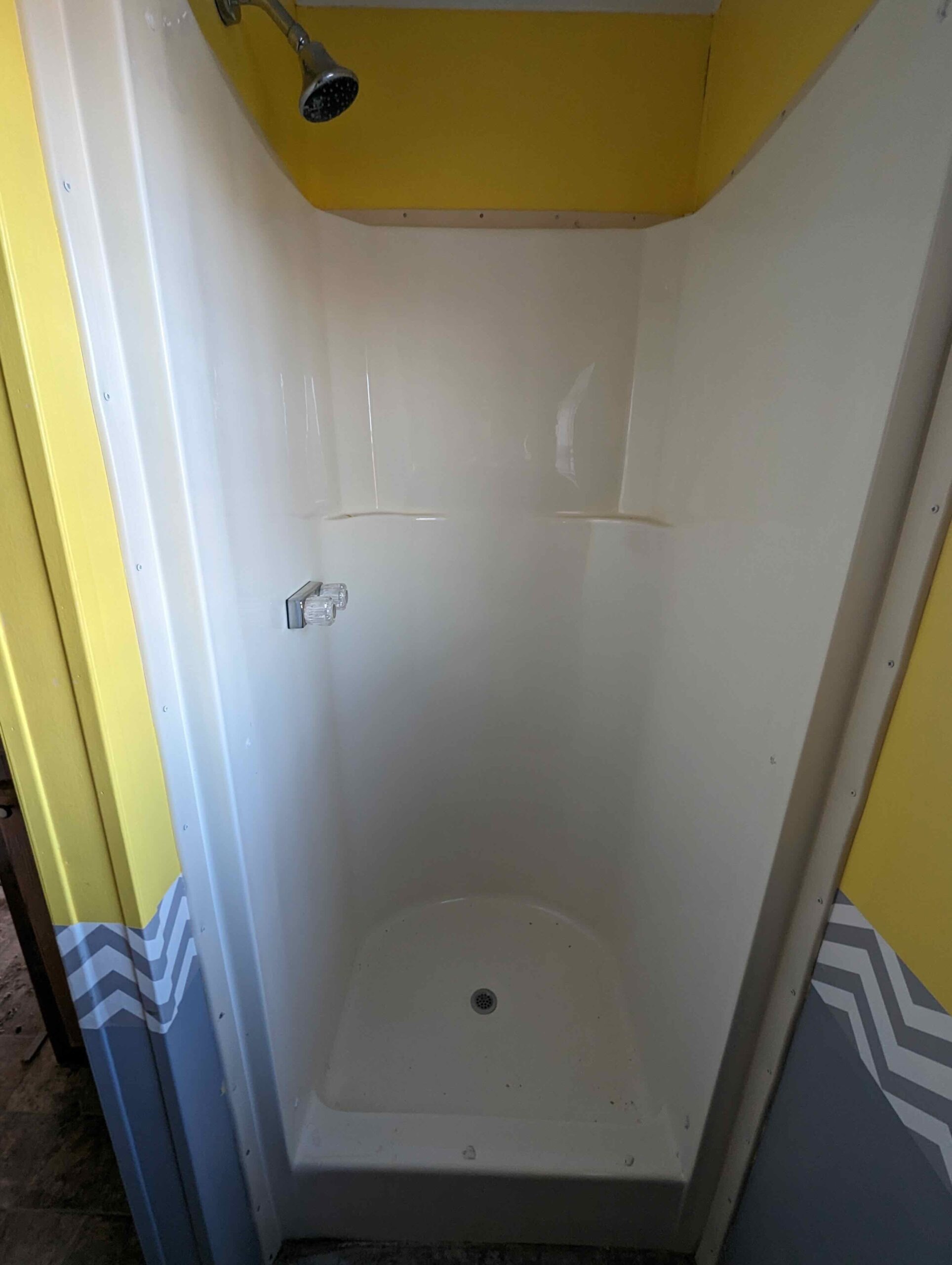 A small, enclosed shower with white walls and a single silver showerhead. The adjacent walls are bright yellow with gray accents, creating a cheerful, clean atmosphere.