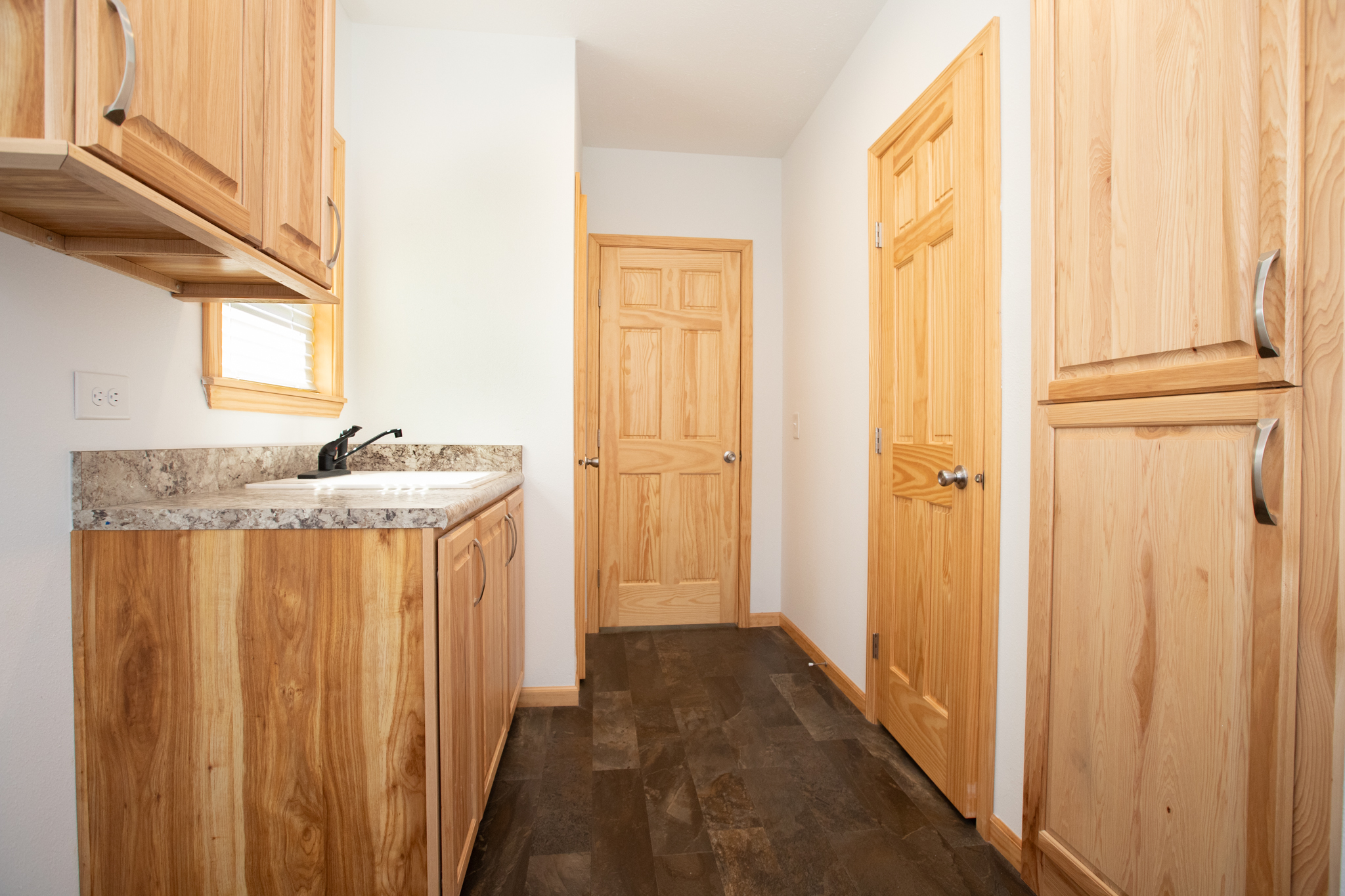 Narrow kitchen with wood cabinets, black faucet, and stone countertop on the left. Wooden door and pantry on the right. Warm, inviting tone.