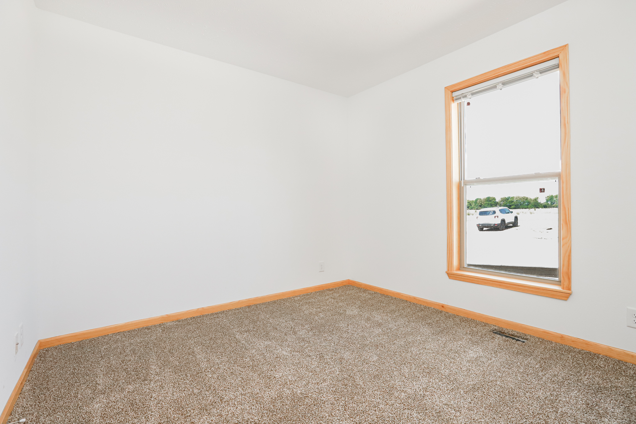 Empty room with white walls and carpeted floor. A single window reveals a sunny view of a parked car and trees outside. Bright, clean, and minimalistic atmosphere.