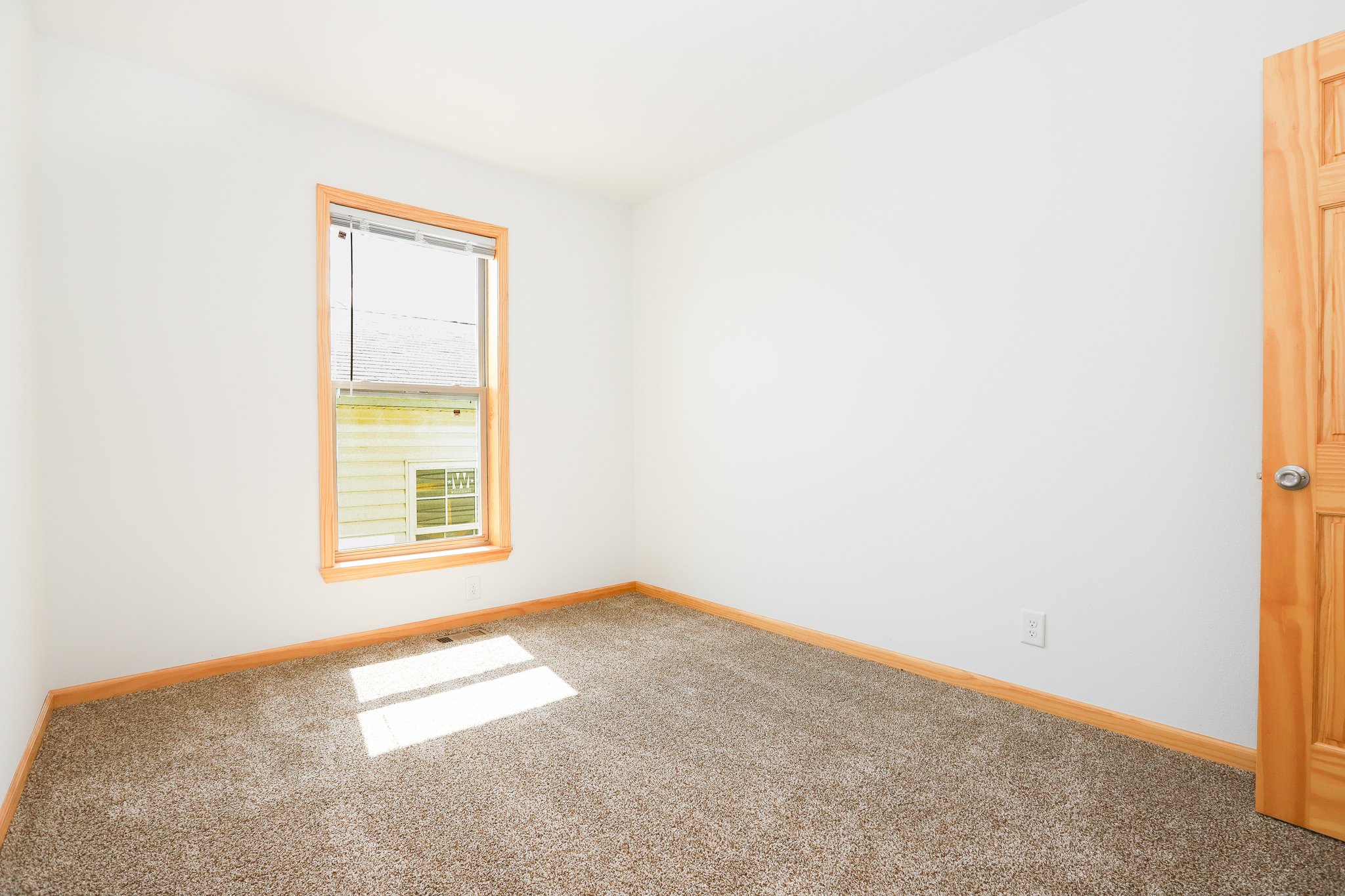 Empty room with white walls, beige carpet, and a light wooden door. Sunlight streams through a window with light wood framing, creating a serene atmosphere.