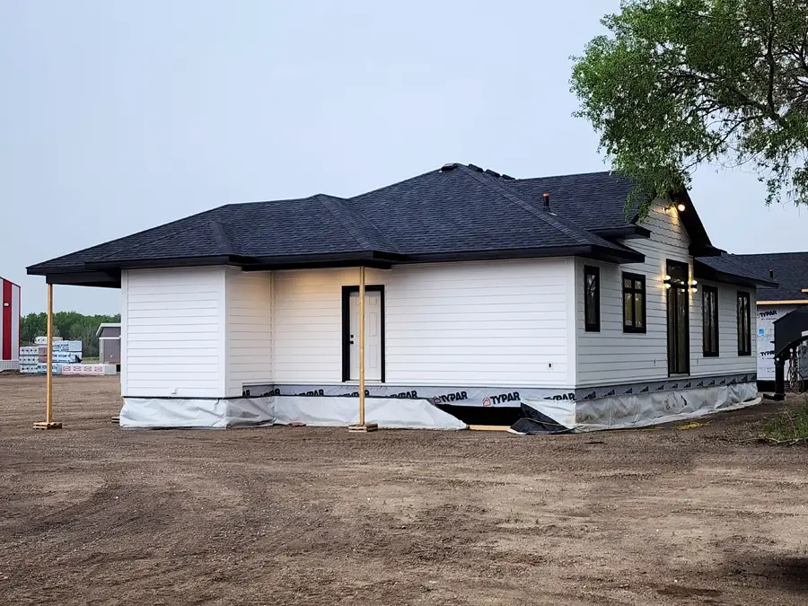 Newly constructed white house with a dark roof, surrounded by a dirt yard. The exterior is unfinished, with visible Typar wrap and support beams.