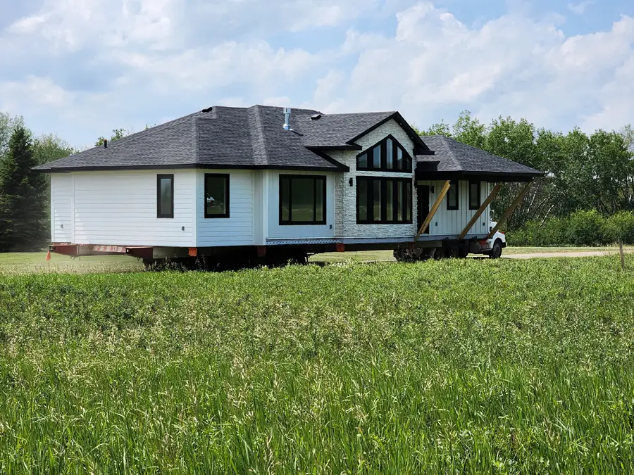 A white house with a black roof, placed on a trailer for transport, sits in a green field. The sky is cloudy, creating a calm and peaceful setting.