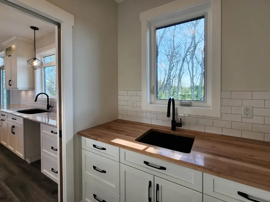 Modern kitchen with light wood countertops and black fixtures. Sunlight streams through a window, highlighting white cabinets and subway tiles. Bright and airy ambiance.