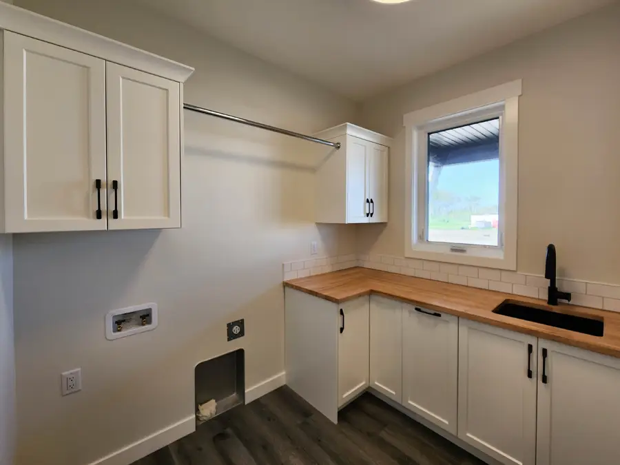 Laundry room with white cabinets, wooden countertop, and black faucet. A clothes rod is above a washer space. Large window lets in natural light.