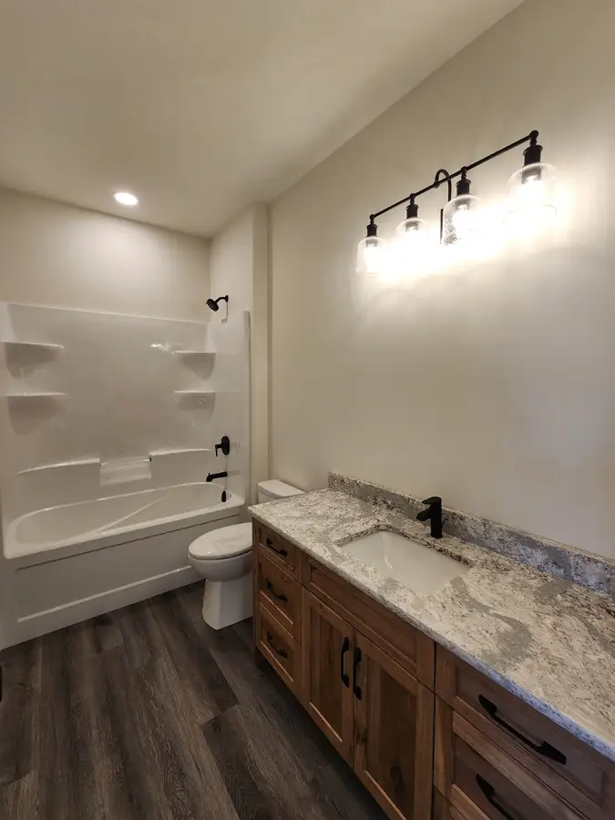 Modern bathroom with wood vanity and granite countertop. Overhead lights illuminate the space. White bathtub with black fixtures on the left.