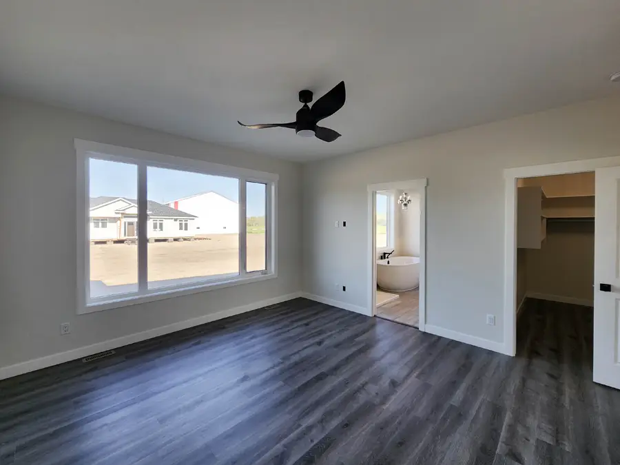 Modern, empty room with dark wood floors and large window showcasing a suburban view. Black ceiling fan and open doors leading to a bathroom with a tub and a closet.