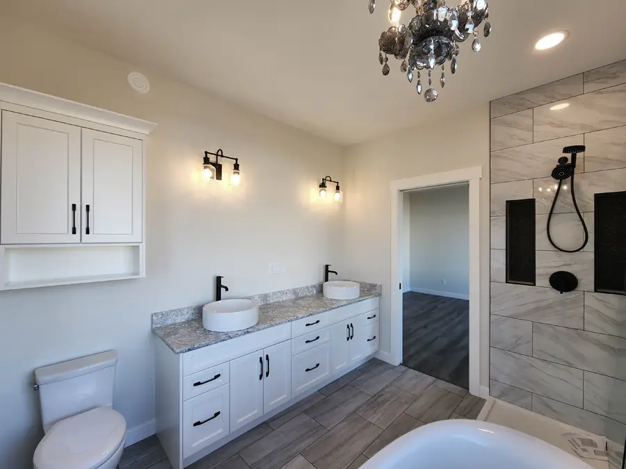 Elegant bathroom with dual sinks on a granite counter, under modern sconces. Visible are a sleek toilet, marble-tiled shower, and a crystal chandelier.