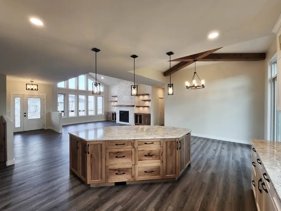 Spacious open-concept living area with wooden floors, a granite-topped kitchen island, modern pendant lights, and beams. Fireplace and large windows in the background.