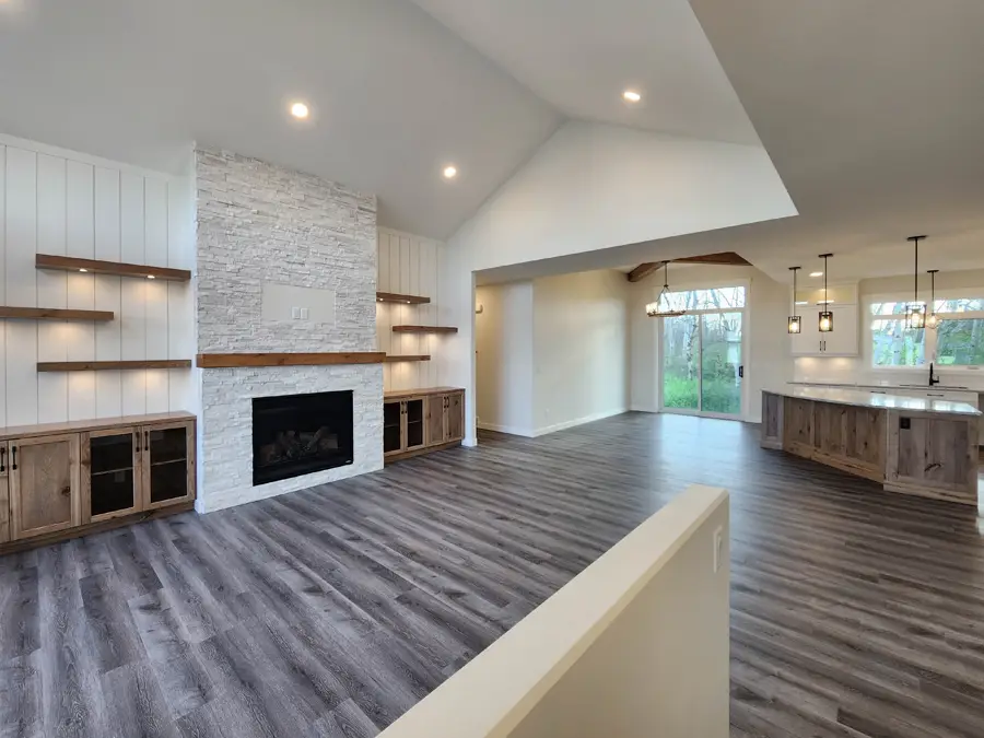 Spacious modern living area with vaulted ceiling. Features a stone fireplace, wooden shelves, dark laminate flooring, and an open kitchen with island.
