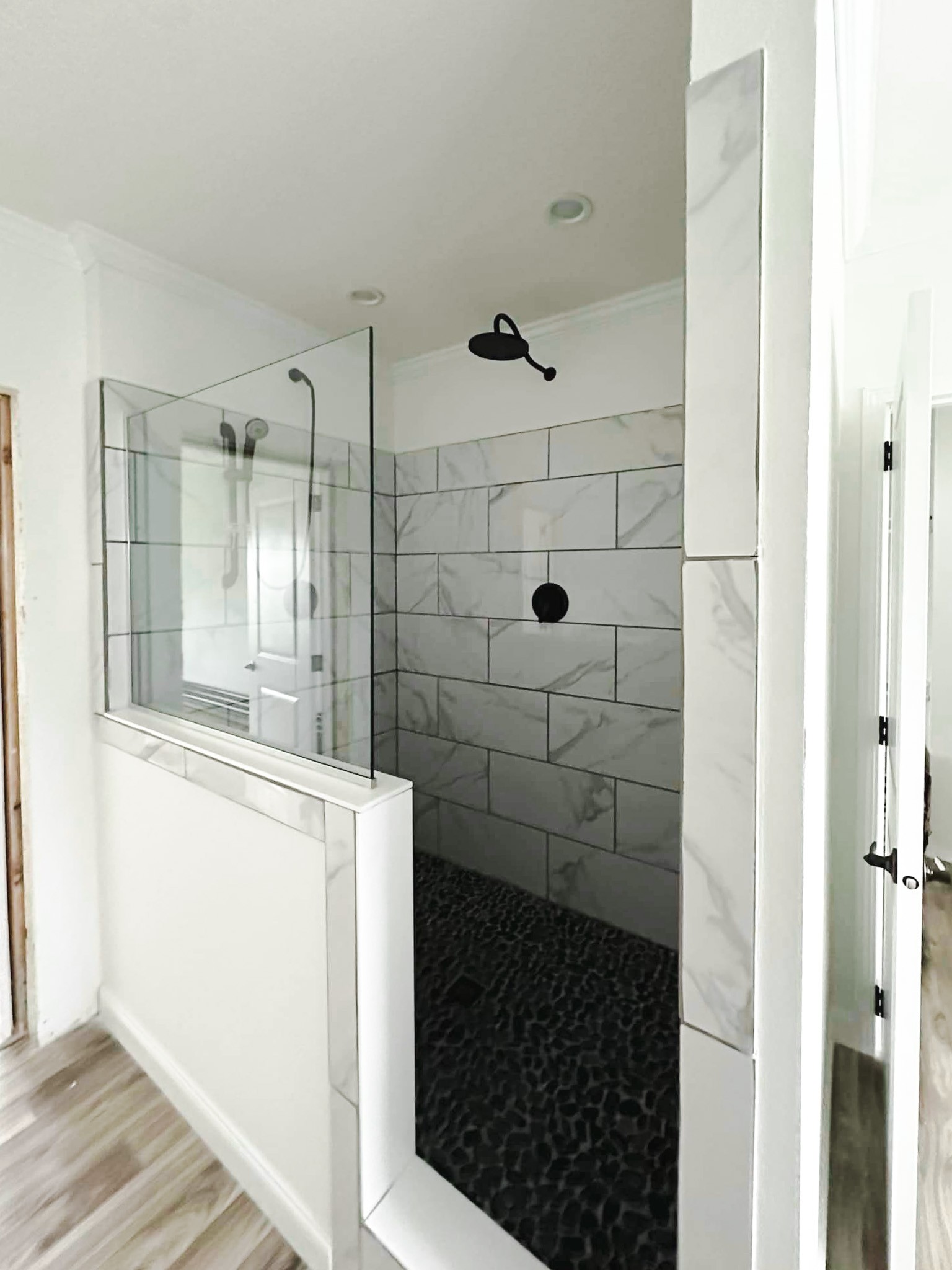 Modern shower with glass walls, gray marbled tiles, and a black showerhead. Pebble-like dark floor contrasts with light wood flooring outside. Sleek and minimalistic design.
