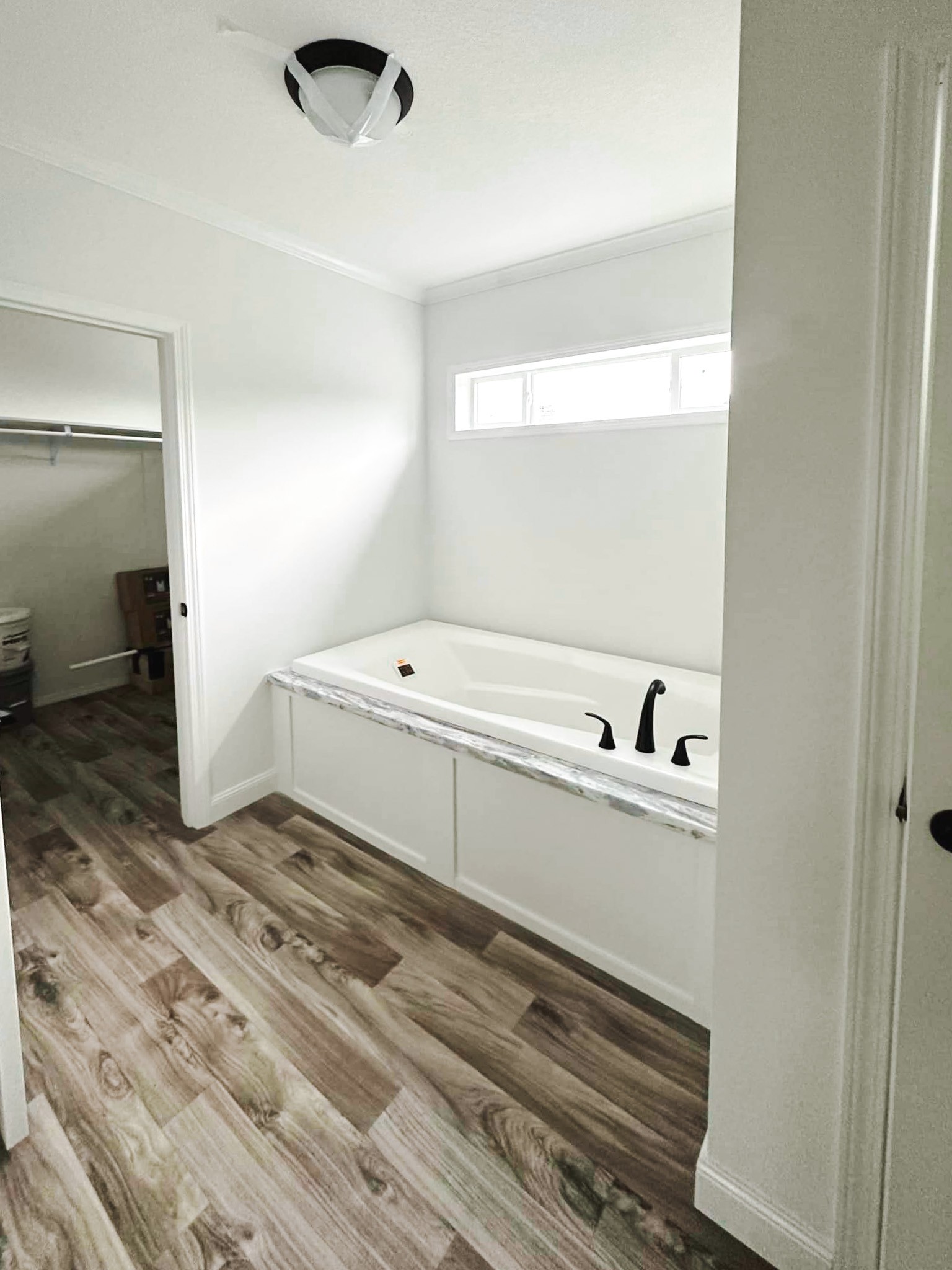 Modern bathroom featuring a white corner bathtub with marble surround and black fixtures. Wood-patterned flooring complements the light ambiance.