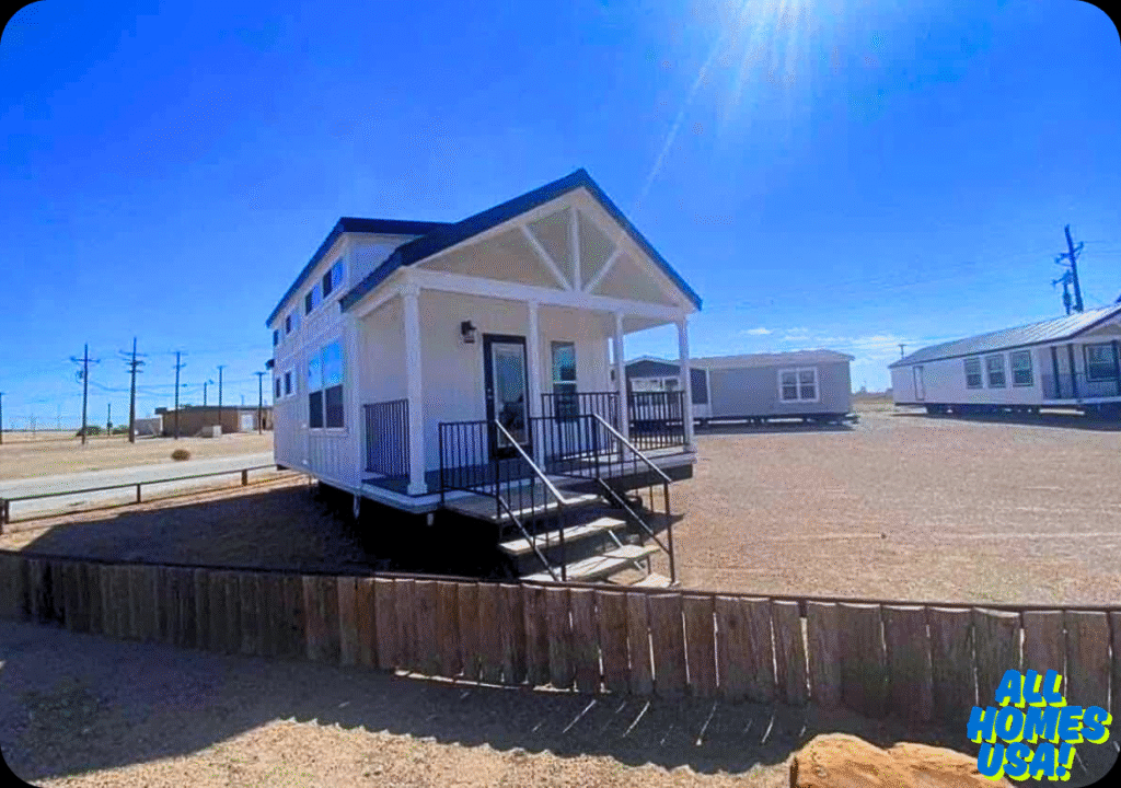 A small, white, modern modular home with a porch and steps sits in a sunny, desert landscape. Nearby, other similar homes dot the barren terrain. Image for illustrative purposes.