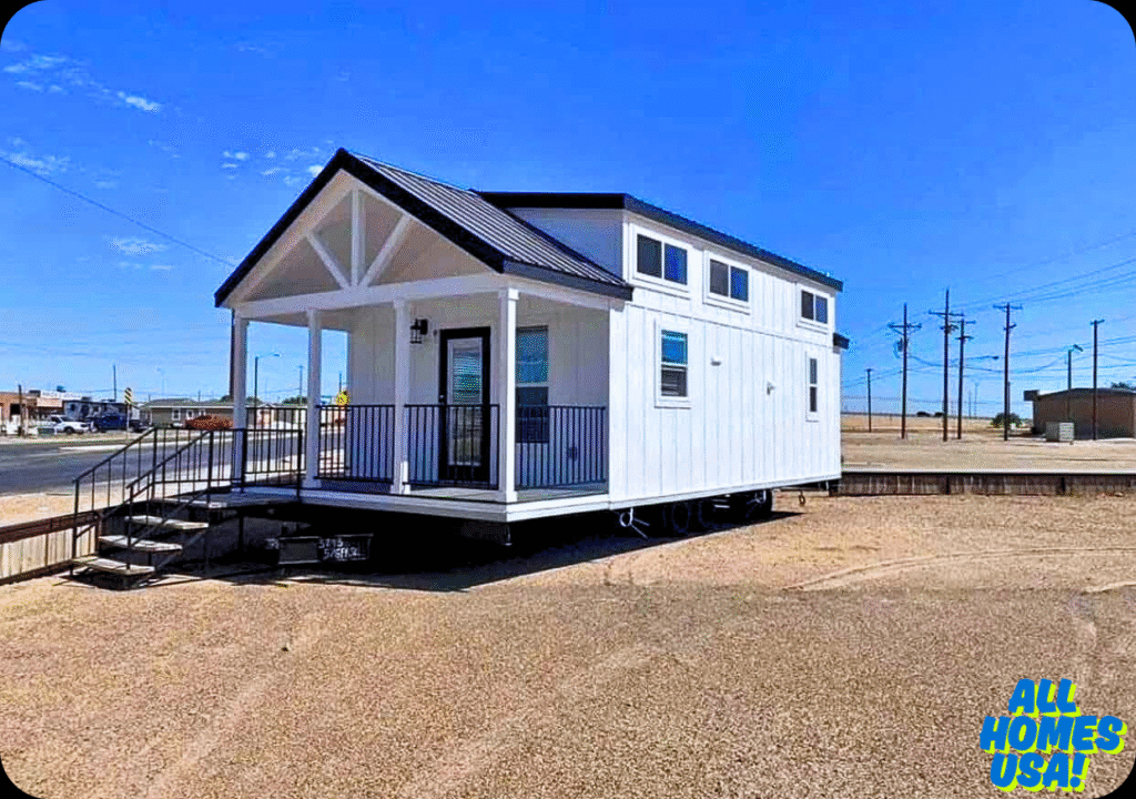 A modern tiny house with white siding and a black roof sits on a sandy lot under a clear blue sky. It features a small porch with black railings. Image for illustrative purposes.