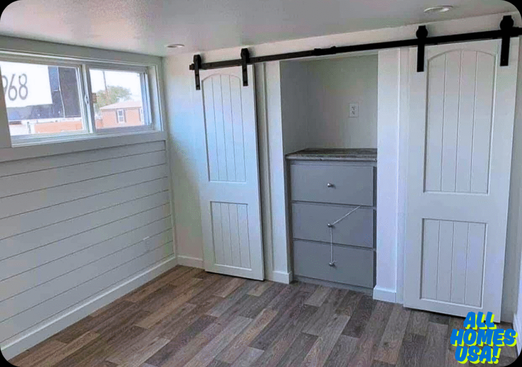 Bright room with light wood flooring, a small closet with sliding barn doors, white shiplap walls, and a three-drawer chest. Logo reads "All Homes USA!" Image for illustrative purposes.