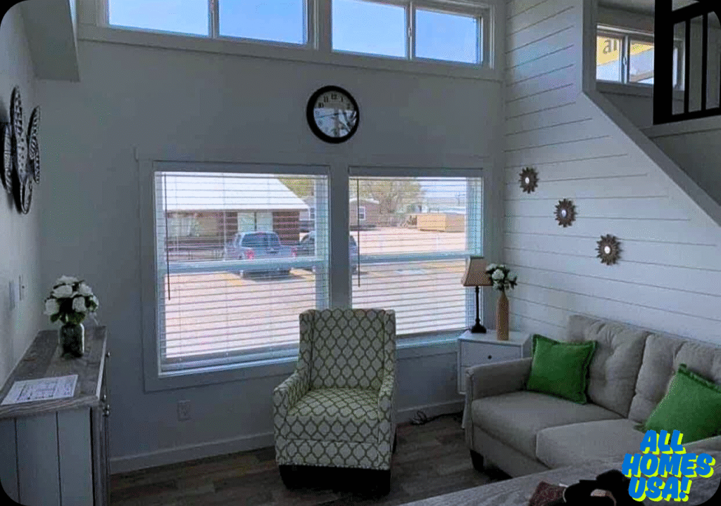 Bright living room with a patterned armchair, beige sofa with green cushions, and decor. Large windows, a wall clock, and flowers create a cozy vibe. Image for illustrative purposes.