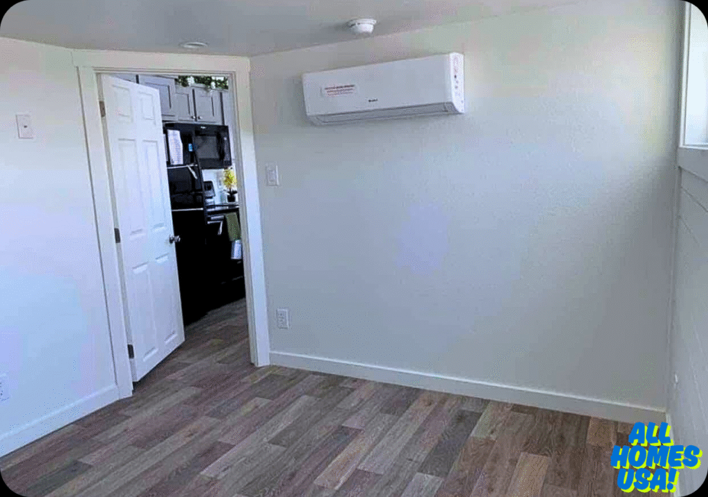 A small, empty room with white walls and wood flooring. An open door reveals a kitchen with dark cabinets. A wall-mounted AC unit is above the door. Bright and minimalistic. Image for illustrative purposes.