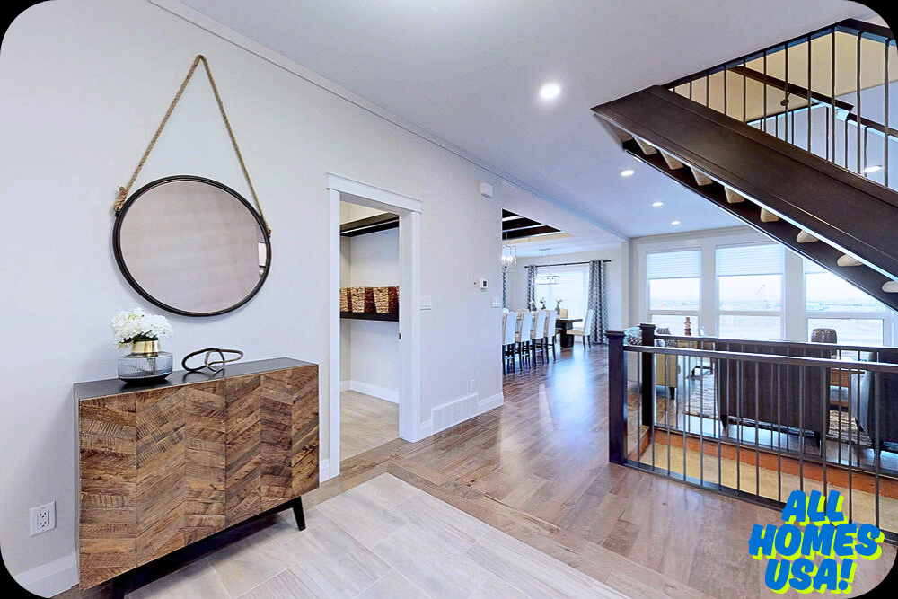 Modern home interior with wooden floors, open staircase, and large windows. A round mirror and wooden console table adorn the entrance. Bright and inviting atmosphere. Logo reads “All Homes USA.” Image for illustrative purpose.