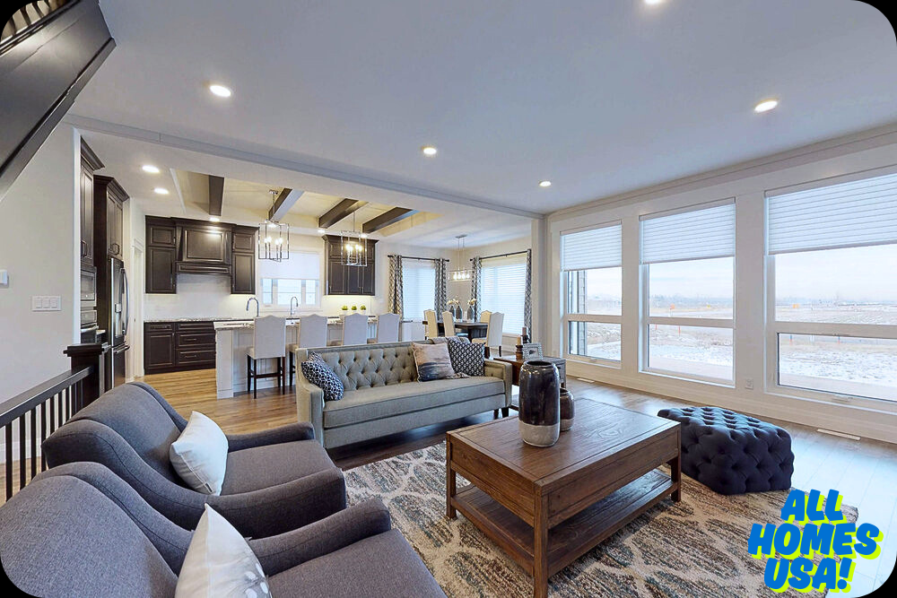 Spacious open-concept living area with a modern kitchen, featuring dark cabinets and a breakfast bar. Cozy seating and decorative items create a welcoming atmosphere. Large windows provide ample natural light. "All Homes USA" logo in the corner. Image for illustrative purpose.