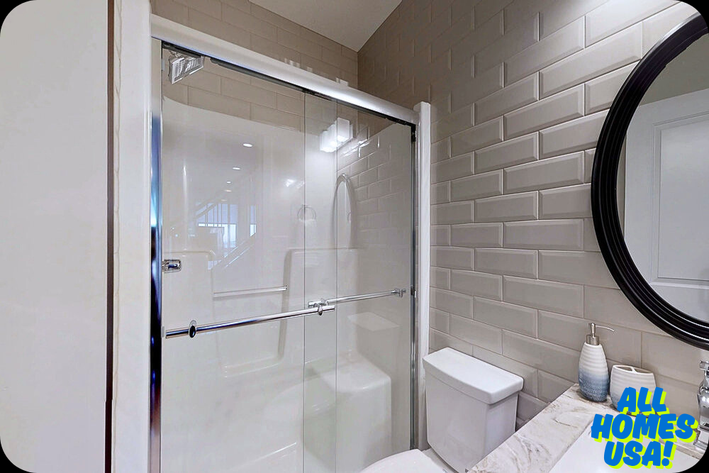 Bright bathroom with beige subway tiles, glass shower doors, round mirror, and countertop with soap dispenser. Modern and clean feel. Logo in corner. Image for illustrative purpose.