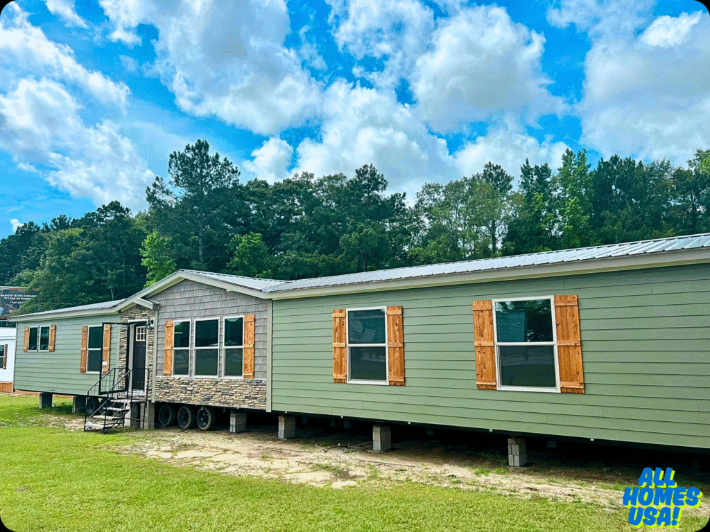 A green manufactured home with wooden shutters and stone accents sits on a lush lawn, bordered by a dense forest and a cloudy blue sky. Image for illustrative purposes.