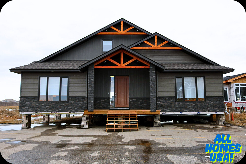 A modern, dark-colored house with wood accents is elevated on blocks and surrounded by a wet, unfinished lot. "ALL HOMES USA!" logo is in the lower right corner. Image for illustrative purpose.