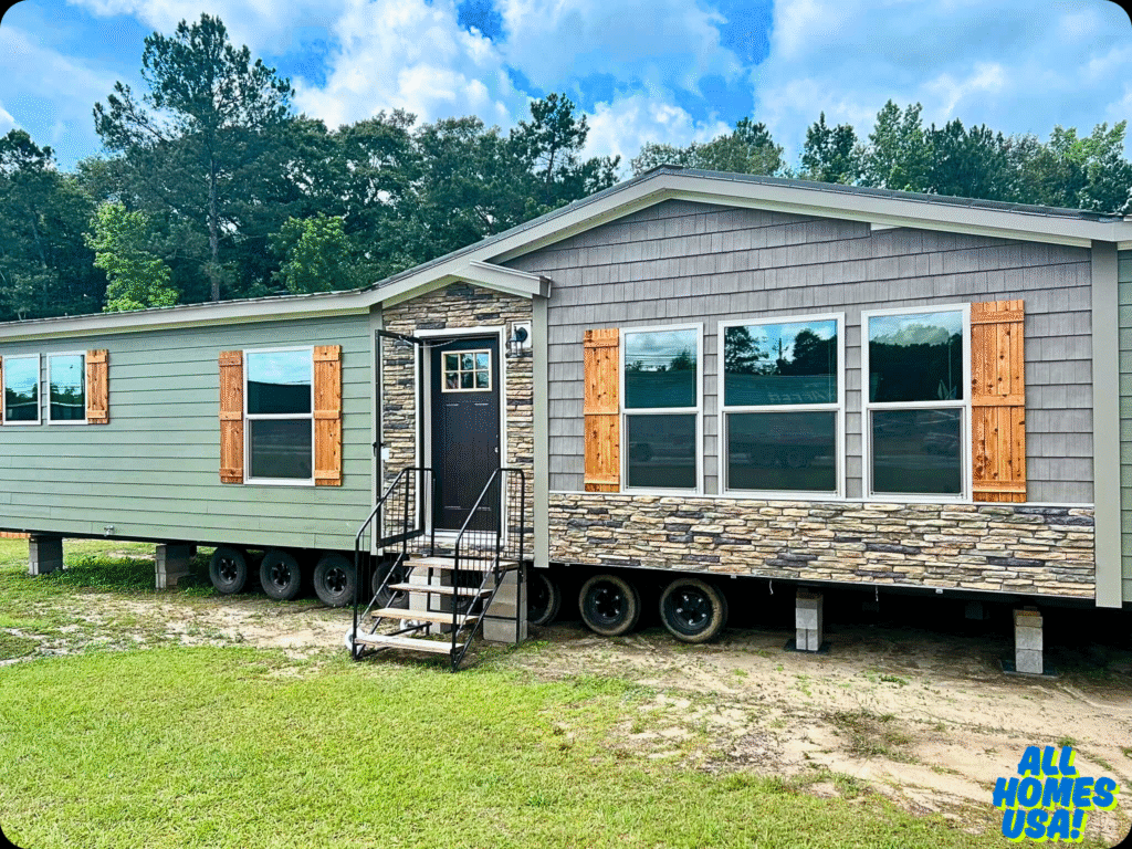 Modular home on wheels with stone and wood siding, large windows, and wooden shutters. It's set in a grassy area with trees in the background. Image for illustrative purposes.