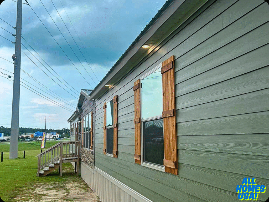 Side view of a green manufactured home with wooden shutters and a small porch. Overcast sky with power lines. A “All Homes USA!” logo in the corner. Image for illustrative purposes.